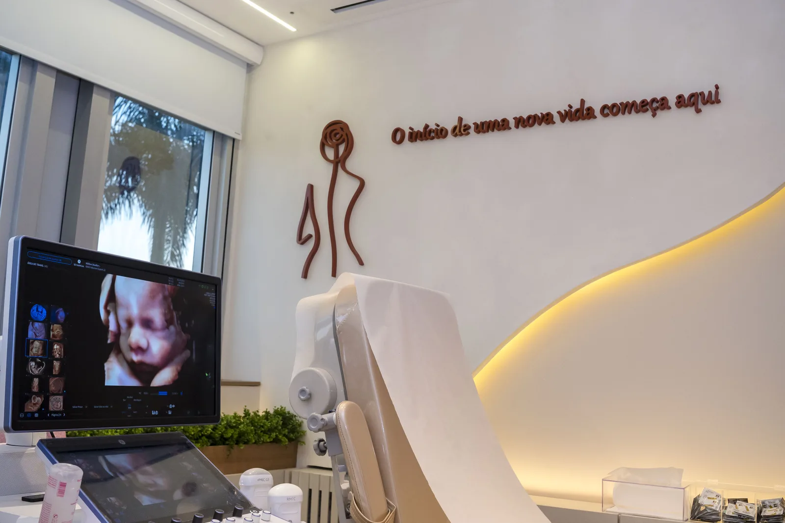 Sala de ultrassonografia do consultório, com monitor de exame e a frase O início de uma nova vida começa aqui aplicada na parede